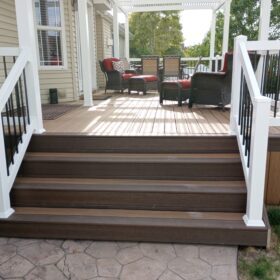 Create the perfect low-maintenance backyard retreat with a custom composite deck from DeckPro LLC. This beautiful project features rich brown composite decking paired with elegant white vinyl railings and contrasting black metal balusters. The attached white pergola provides comfortable shade, while the wide stairs offer an inviting transition to the stamped concrete patio below.