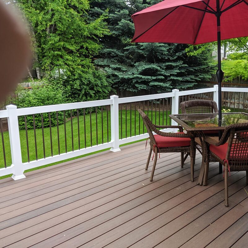 An elevated view across a newly installed multi-tonal composite deck surface, showcasing a comprehensive outdoor living solution by DeckPro LLC. The deck features grooved composite planks in warm brown and grey tones. Centered on the deck is a stylish dark brown wicker dining set for four, featuring a round glass-top table and four armchairs outfitted with vibrant red seat and back cushions.