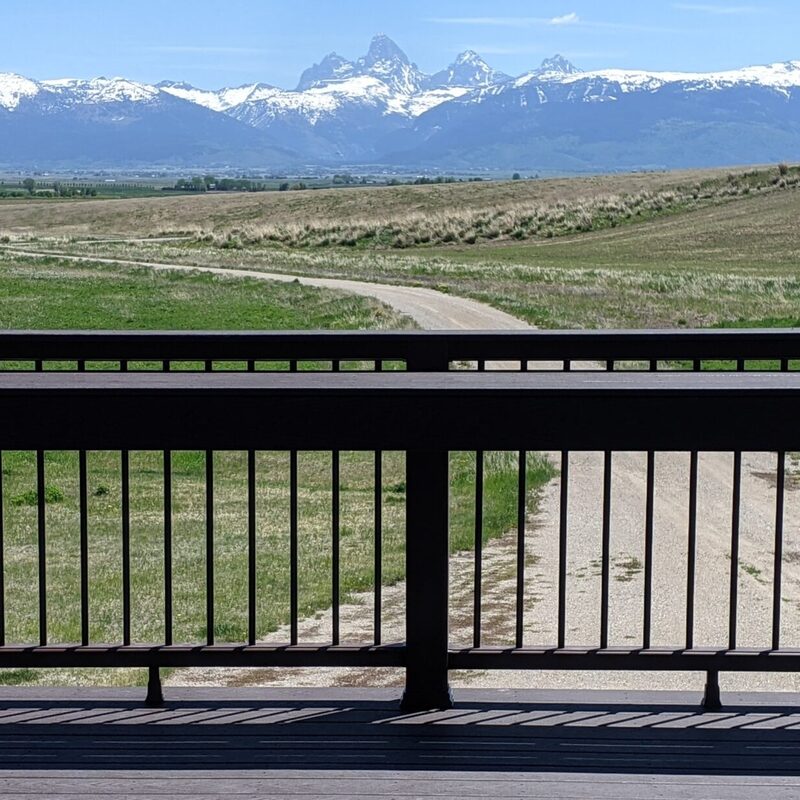 Expand your outdoor living space with this cozy raised deck by DeckPro LLC. We installed high-quality, low-maintenance composite decking in a warm brown tone and paired it with safe and sophisticated dark vertical perimeter railings, creating a sturdy retreat perfect for relaxation. Pictured on a beautiful sunny day with clear skies and distinct shadows cast across the deck boards, this functional build transitions through a massive and inviting staircase with matching composite planks leading down to a lower patio level, creating defined sun and shade areas and optimizing views of the vast backyard. Direct access is available via a sliding glass door.