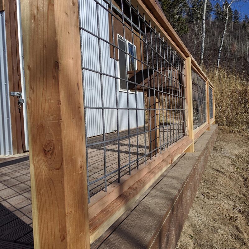 A durable and stylish deck on a mountain property featuring a unique railing system that combines rustic wood posts with modern wire mesh panels. The mesh panels offer unobstructed views of the surrounding natural landscape, including the evergreen trees and dried grass. This design provides both safety and a visual connection to the mountain environment.