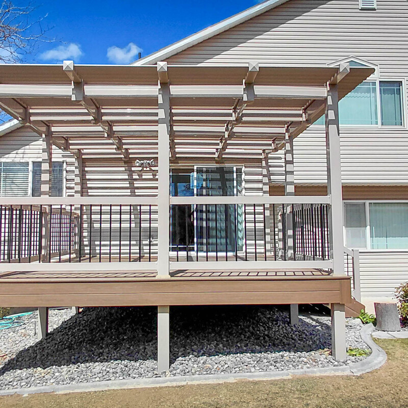 Exterior wide shot of a tan elevated composite deck with an attached pergola, black metal railings, and side stairs against a light-colored siding house.

