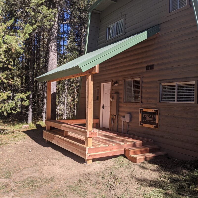 Extend your multi-directional natural views with this rustic custom composite deck and front porch addition by DeckPro LLC. We took a classic mountain log cabin home and created a sophisticated and sturdy functional retreat, pictured bordering a dirt road/trail in natural vegetation. Pictured on a beautiful sunny day, this unique tranquillity sanctuary features rich brown wood-grain composite decking planks with a defined clean picture frame board pattern around the perimeter for a safe and sophisticated finish.