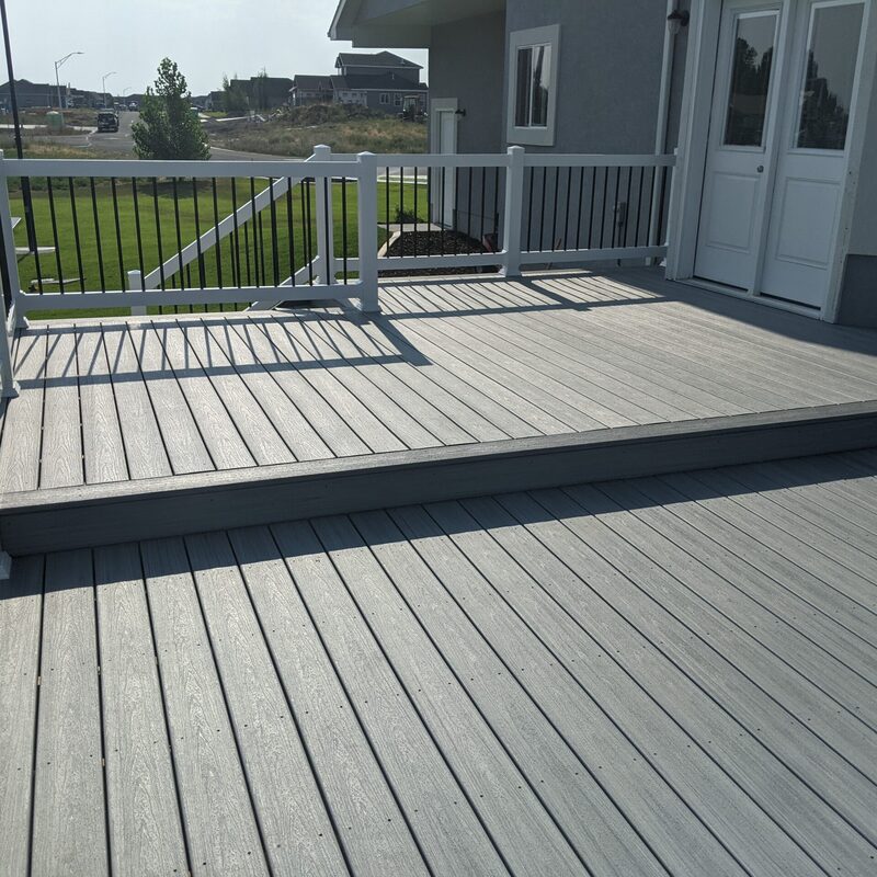 Expand your upper-level living space with this cozy raised fir balcony deck by DeckPro LLC, which combines rustic elements with modern comfort. This sturdy and stylish retreat uses extensive natural wood decking planks and a full perimeter railing system with wide top rails to create a safe, open retreat. Pictured on a sunny day with distinctive railing shadows, the build overlooks a natural dirt road, expansive pine trees, and a distant basketball hoop, creating a tranquil mountain retreat perfect for taking in scenic woodland views.