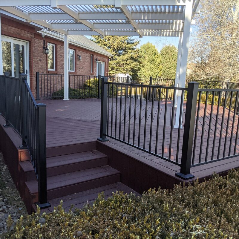 This elegant project by DeckPro LLC features a rich red composite deck that perfectly complements the home's brickwork. The white vinyl pergola provides beautiful filtered shade, while the black metal railing adds a modern touch to this backyard retreat.