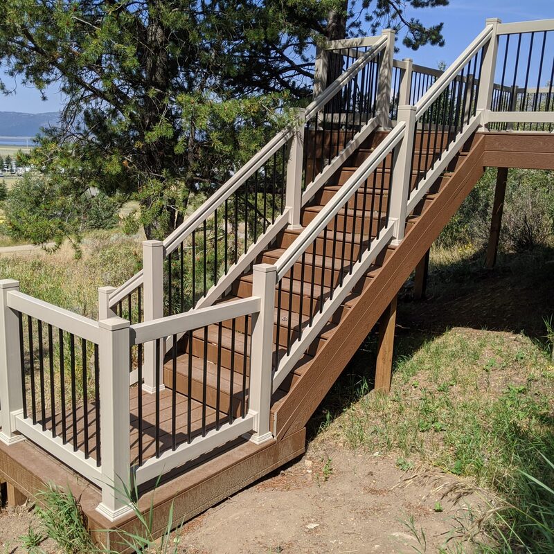A steep, multi-level set of composite deck stairs ascending a hillside, featuring a sophisticated hybrid railing system. The stairs are constructed from a durable, rich brown composite decking (AZEK style) for a maintenance-free wood look. The hybrid railing combines sturdy, taupe-colored composite posts and top/bottom rails with contrasting dark, round vertical aluminum balusters (black finish) for safety and a sleek aesthetic.