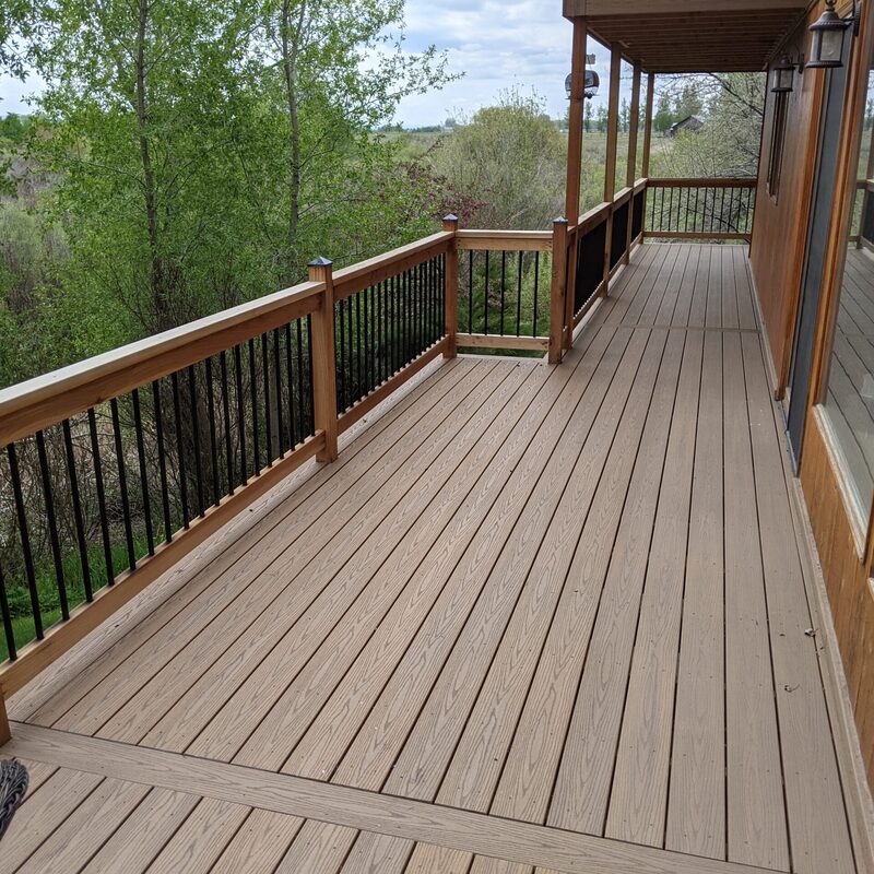 Enhance your home's value and outdoor lifestyle with our Rustic Riverview Custom Deck & Hybrid Railing Project by DeckPro LLC. This massive, covered composite deck spans the length of the building, providing a durable and sophisticated outdoor living space with breathtaking, expansive views of a wooded river valley. We custom-engineered a hybrid railing system, blending warm, stained wood posts and top/bottom rails with sleek, black metal balusters (pickets) and pyramid post caps. The durable, grooved low-maintenance composite decking surface, complete with an elegant solid-plank border, seamlessly integrates indoor and outdoor spaces through large floor-to-ceiling windows and sliding glass doors. The covered design ensures all-weather functionality, making this the perfect retreat for relaxing and entertaining. Trust DeckPro LLC for your custom, high-end deck designs and installations.