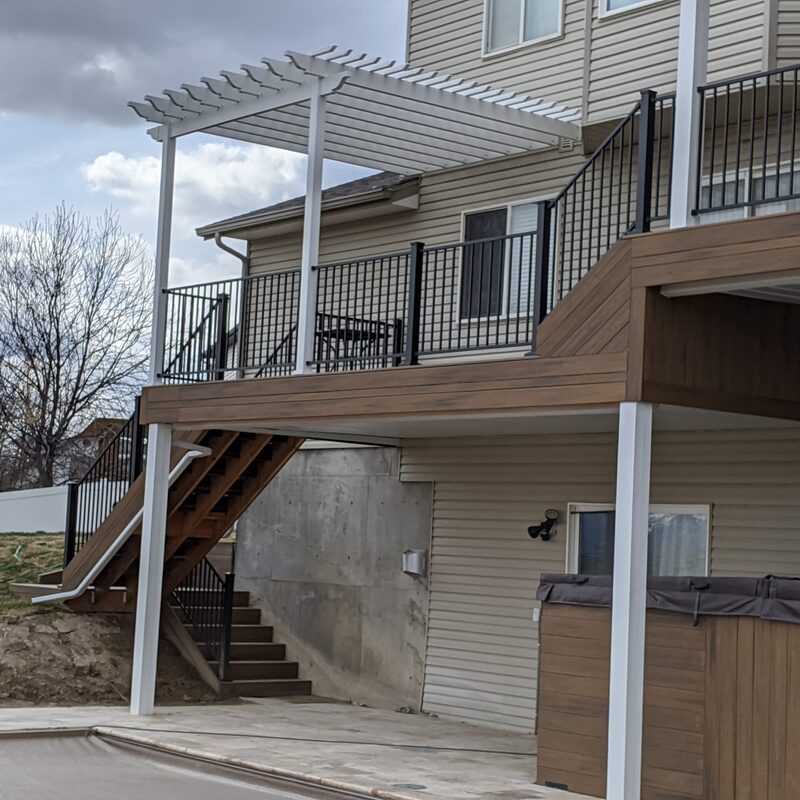 Extend your multi-level outdoor living with this expansive composite deck and patio transformation by DeckPro LLC. We took a multi-story home with rough grading and rough landscaping, pictured bordering natural vegetation, and turned it into a sophisticated, multi-functional sanctuary perfect for large-scale entertaining or tranquil relaxation. Pictured on a beautiful day with distinct shadows cast across the planks, this massive structure features expansive gray composite planks paired with sleek black metal perimeter railings