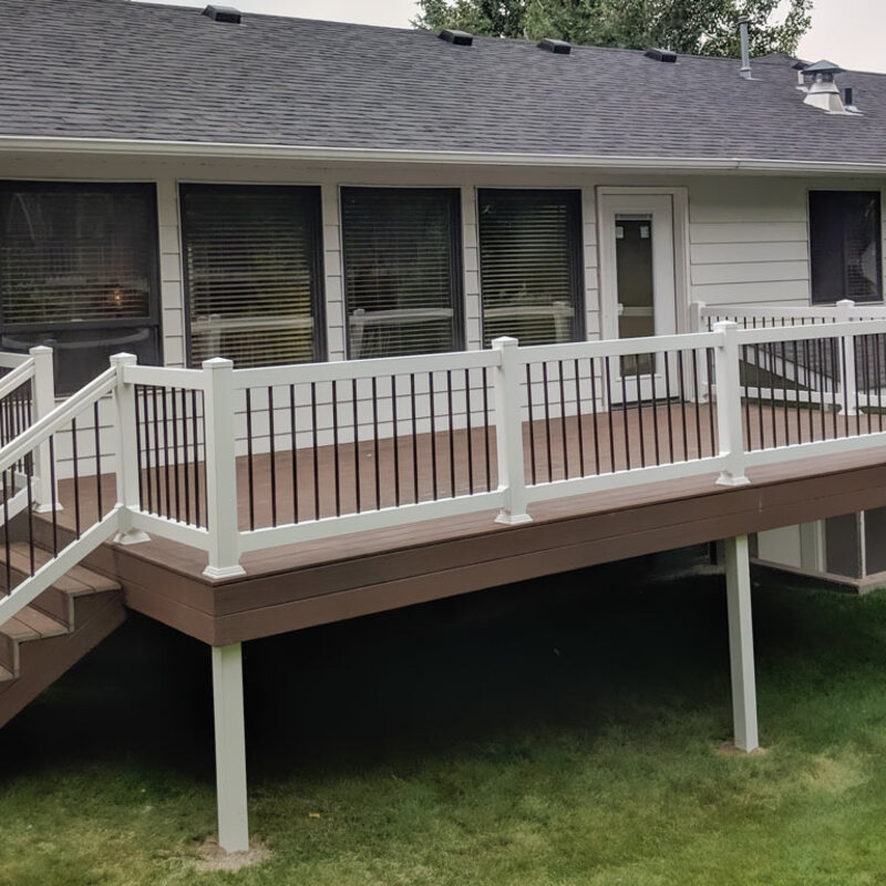 Extend your upper-level living space with this cozy raised balcony deck by DeckPro LLC. We installed durable, low-maintenance composite decking in a warm brown tone that transitions seamlessly from the rustic log cabin architecture. Pictured on a sunny day with distinctive railing shadows, this custom build features sleek black metal perimeter railings to ensure unobstructed woodland views, with direct access via a white-trimmed glass door.