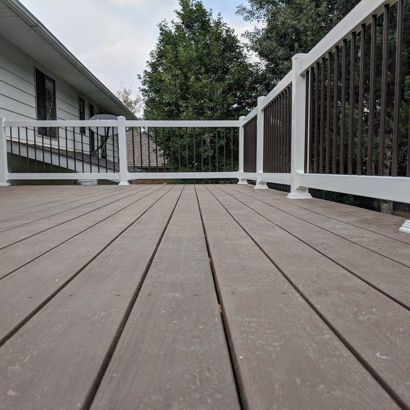 This project features a stunning elevated composite deck with integrated railings. The use of a rich brown composite decking offers the look of wood with significantly less maintenance. The perimeter is secured by durable white composite posts and railings, accented with contemporary dark vertical pickets, creating a safe and stylish outdoor living space. The design includes a perimeter gate leading to a staircase, attached seamlessly to the home’s existing siding and sliding door.